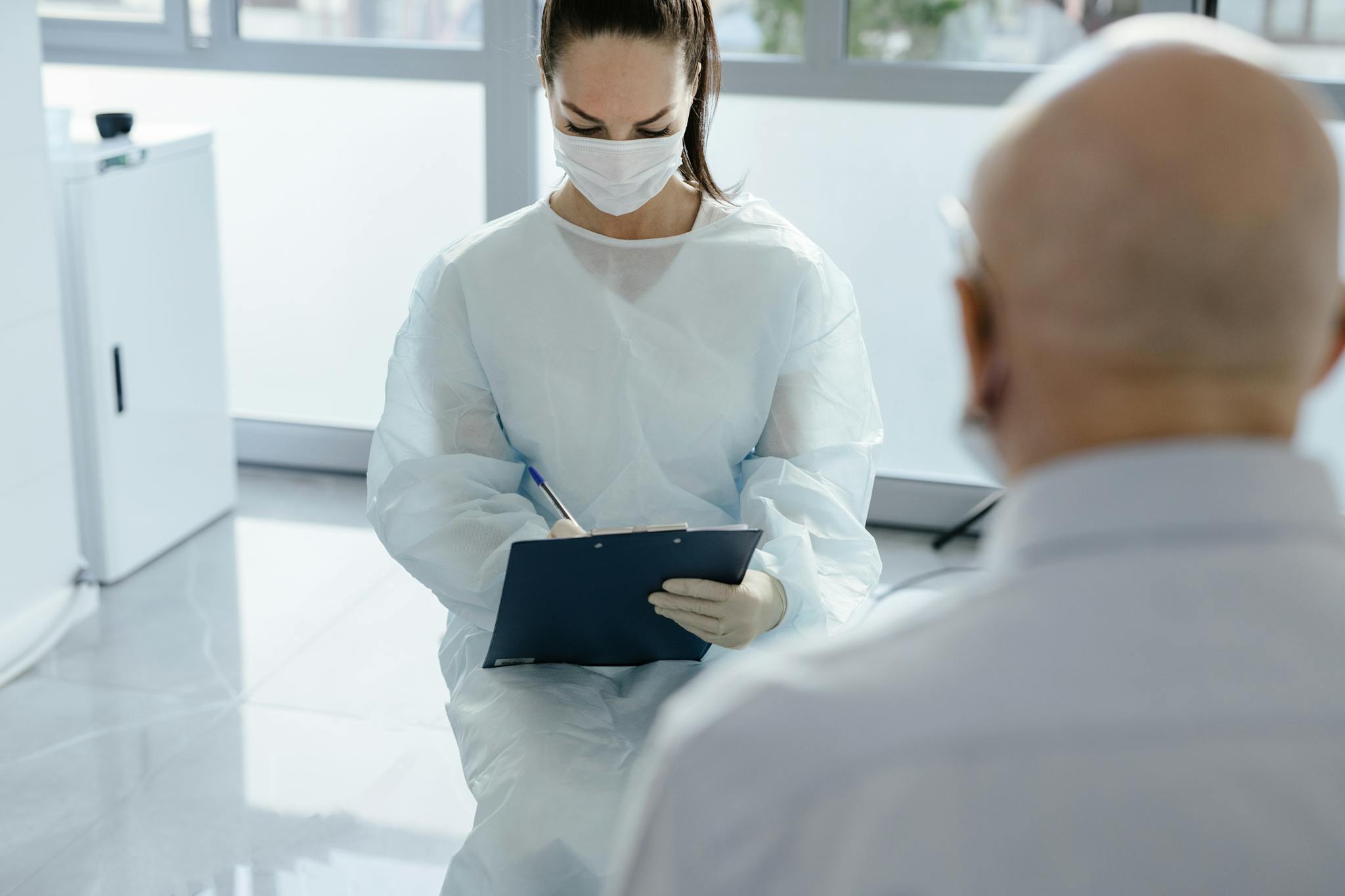 Doctor in protective gear taking notes during a medical consultation with a patient.
